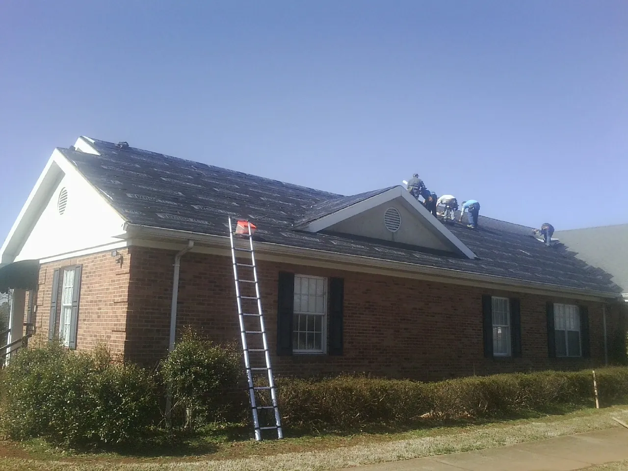 Crew tearing shingles off a brick ranch roof with a ladder set — Metal Roof Repair work by our crew in Sterrett