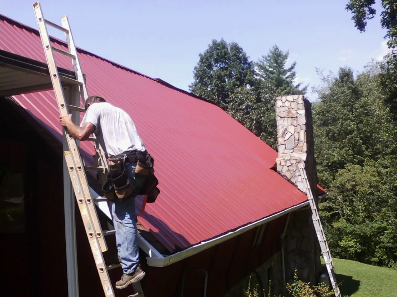Roofer on a ladder beside a red metal roof and stone chimney — Soffit & Fascia Repair by Roofing Brothers in Sterrett